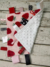 Load image into Gallery viewer, A strawberry-themed sensory baby lovey, featuring a pink fabric cover with red strawberries, a white textured minky dot back, and multiple colorful ribbon tabs, displayed on a rustic white-washed wooden background.
