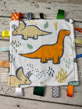 Load image into Gallery viewer, A square white plush baby sensory cloth featuring cartoon dinosaurs in orange, beige, and green, decorated with colorful ribbon tabs on a rustic wooden background.
