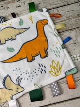 Load image into Gallery viewer, A soft plush sensory blanket featuring cartoon dinosaurs and colorful ribbon tabs, laid flat on a rustic wooden surface.
