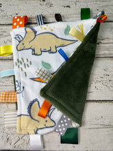 Load image into Gallery viewer, A baby sensory lovey blanket featuring a white front with a dinosaur print, a dark green plush back, and colorful ribbon tabs, displayed on a rustic white wood background.
