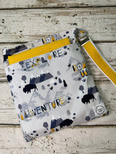 Load image into Gallery viewer, A fabric pouch with a western-themed print featuring bison, mountains, and the words 'EXPLORE' and 'ADVENTURE' in yellow, navy, and gray. The pouch has a yellow and white polka dot strap with a silver metal clasp, laid flat on a rustic white-washed wooden background.
