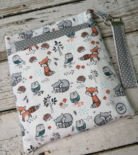 Load image into Gallery viewer, A fabric pouch with a woodland animal print featuring foxes, owls, raccoons, and hedgehogs on a white background, with gray polka dot trim and a matching wrist strap, displayed on a rustic white-washed wooden surface.

