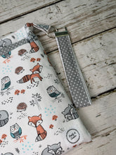 Load image into Gallery viewer, A fabric pouch with a woodland animal print featuring foxes, owls, and hedgehogs on a white background, complete with a gray polka dot wrist strap and metal key ring, displayed on a rustic white wooden surface.
