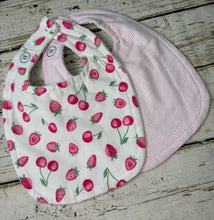 Load image into Gallery viewer, A two-piece baby bib set on a rustic white-washed wooden background. The front bib is white with a pink strawberry and cherry print, and the back bib is light pink with thin white stripes.
