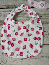Load image into Gallery viewer, A white fabric baby bib with a repeating strawberry print, laid flat on a rustic whitewashed wooden surface. A second striped bib is partially visible in the top right corner.
