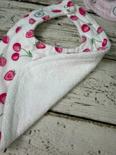 Load image into Gallery viewer, A close-up of a baby bib with a white front featuring a pink strawberry and cherry print, and a soft white terrycloth backing, laid on a distressed white wooden surface.
