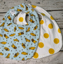 Load image into Gallery viewer, Two fabric baby bibs laid flat on a rustic whitewashed wood surface. The front bib is light blue with a repeating print of yellow Winnie the Pooh print.  The back bib is white with large yellow dots.

