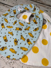 Load image into Gallery viewer, Two fabric baby bibs laid flat on a rustic whitewashed wood surface. The front bib is light blue with a repeating print of yellow Winnie the Pooh print.  The back bib is white with large yellow dots.
