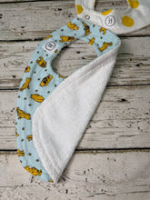 Load image into Gallery viewer, A baby bib laid flat on a rustic white-washed wood surface. The bib has a light blue Winnie the Pooh print and shows the terrycloth white back
