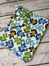 Load image into Gallery viewer, A folded baby blanket and knit hat featuring a colorful cartoon monkey print on a white background, styled on a rustic whitewashed wooden surface.
