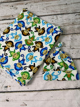 Load image into Gallery viewer, A baby swaddle blanket &amp; hat set featuring a white fabric blanket and matching hat with a colorful cartoon monkey print, displayed on a rustic white-washed wooden surface.
