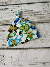 Load image into Gallery viewer, A baby hat with a knotted top, featuring a colorful cartoon monkey print on a white background, laid flat on a rustic white-washed wooden surface.
