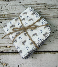Load image into Gallery viewer, A folded white muslin washcloth set of 3 with a blue and gray pattern of small bunnies and leaves, tied with a rustic jute twine bow, placed on a distressed white wooden surface.
