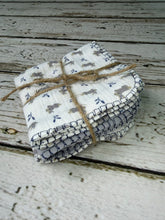 Load image into Gallery viewer, A stack of folded white cotton muslin cloths with a blue and gray woodland pattern, tied with natural jute twine, displayed on a rustic white-washed wooden surface.
