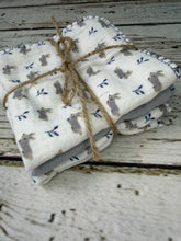Load image into Gallery viewer, A folded stack of white muslin baby blankets with a gray and navy bunny print, tied with natural jute twine, set on a rustic white-washed wooden surface.
