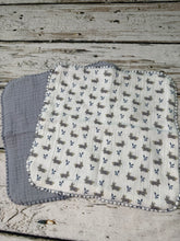 Load image into Gallery viewer, A folded white muslin washcloth set of 3 with a blue and gray pattern of small bunnies and leaves, tied with a rustic jute twine bow, placed on a distressed white wooden surface.
