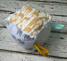 Load image into Gallery viewer, A square baby sensory toy cube with fabric panels featuring giraffe and elephant patterns, textured white dots, and two hanging loops in yellow and mint green, resting on a rustic white wooden plank surface.
