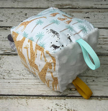 Load image into Gallery viewer, A square baby sensory toy with a white minky fabric body, printed with giraffes, zebras, and palm trees in beige, black, and light blue. It features a mint green ribbon pull tab and a yellow loop on the side, resting on a rustic white wooden plank surface.
