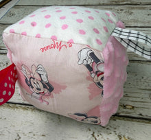 Load image into Gallery viewer, A handmade baby sensory toy cube featuring Minnie Mouse  prints, pink and black polka dot panels, and a white hanging ribbon, resting on a distressed white wooden surface.
