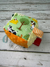 Load image into Gallery viewer, A soft fabric sensory cube toy with a farm theme, featuring a light green front panel with a cow, tree, and goat illustration, orange polka-dot side panels, a yellow-and-white gingham ribbon tab, and a red fabric tag, photographed on a rustic white-painted wood surface.
