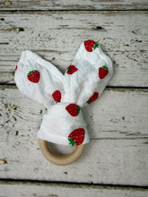 Load image into Gallery viewer, A baby teething toy featuring a white fabric printed with red strawberries and a natural wooden ring, displayed on a rustic white-washed wood background.
