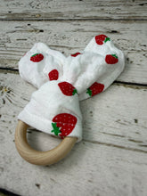 Load image into Gallery viewer, A baby teether toy made from a white muslin cloth printed with red strawberries and green leaves, attached to a natural wooden ring, displayed on a rustic white-washed wooden surface.
