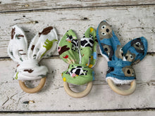 Load image into Gallery viewer, Three wooden teething rings with fabric bunny shaped in different animal prints (dinosaurs, cows, bears) arranged on a rustic white-washed wooden background.
