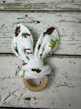 Load image into Gallery viewer, A baby teething ring featuring a white fabric with brown and green dinosaur prints, attached to a natural wooden ring, displayed on a rustic white-washed wood background.
