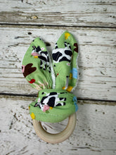 Load image into Gallery viewer, A baby teething ring toy featuring a light green fabric with farm animal cow print  attached to a natural wooden ring, displayed on a rustic white-washed wood background.
