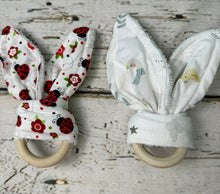 Load image into Gallery viewer, Two fabric baby teething rings with bunny ears displayed on a rustic white-washed wooden surface. The left teething ring has white bunny ears with a red ladybug and floral pattern, and the right one has white bunny ears with a light blue and yellow mother goose pattern.
