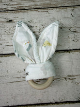 Load image into Gallery viewer, A baby teething ring featuring soft white fabric bunny ears with a small bird and duck print, attached to a natural light wood ring, set against a rustic white-washed wooden background.
