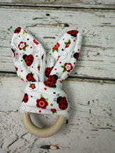 Load image into Gallery viewer, A fabric baby teether with bunny ears and a wooden ring, featuring a white background printed with red flowers, black ladybugs, and green leaves, displayed on a rustic white-washed wooden surface.
