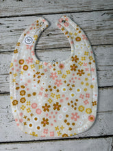 Load image into Gallery viewer, A fabric baby bib with a retro floral pattern in pink, mustard yellow, and brown on a cream background, laid flat on a rustic gray wooden surface.

