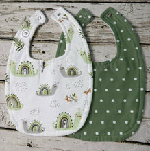 Load image into Gallery viewer, Two baby bibs laid flat on a rustic whitewashed wood surface. The front bib is white with a green and gray rainbow and snail print, and the back bib is olive green with white polka dots.
