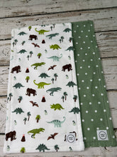 Load image into Gallery viewer, Two baby burp cloths laid flat on a rustic whitewashed wood surface. The front blanket is white with an all-over dinosaur and tree print in green, brown, and gray.
