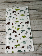 Load image into Gallery viewer, A rectangular burp cloth with a colorful dinosaur and palm tree pattern, laid flat on a rustic, weathered gray wooden surface.
