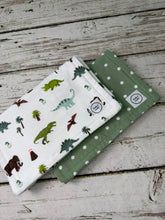 Load image into Gallery viewer, Two folded baby burp cloths displayed on a rustic white-washed wooden surface. The top one is white with a colorful dinosaur and palm tree print, and the bottom is sage green with white polka dots.

