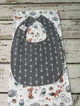 Load image into Gallery viewer, A dark gray baby bib with a white arrow pattern, laid flat on top of a white burp cloth featuring woodland animal prints like foxes, owls, and hedgehogs, set on a rustic wooden surface.
