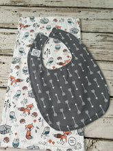 Load image into Gallery viewer, A dark gray baby bib with a white arrow pattern, laid flat on top of a white burp cloth featuring woodland animal prints like foxes, owls, and hedgehogs, set on a rustic wooden surface.
