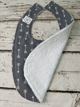 Load image into Gallery viewer, A baby bib with a dark gray fabric printed with white arrows, featuring a soft white terrycloth lining, laid flat on a rustic white-washed wooden surface.
