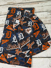 Load image into Gallery viewer, A set of Detroit Tigers themed baby burp cloth and bib featuring an all-over graphic print of the team logo, baseballs, and 'Detroit Tigers' script on a navy blue background, laid flat on a white-washed wooden surface.
