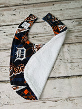 Load image into Gallery viewer, A baby bib with a Detroit Tigers baseball print on the outside and a soft white terrycloth lining, laid flat on a rustic white-washed wooden surface.
