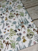 Load image into Gallery viewer, A burp cloth with a Bambi woodland animal print, featuring deer, rabbits, squirrels, and butterflies among trees and flowers on a white background, laid flat on a rustic wooden surface.
