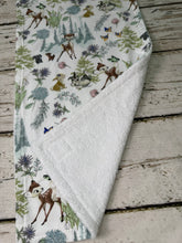 Load image into Gallery viewer, A burp cloth and bib with a Bambi woodland animal print, featuring deer, rabbits, squirrels, and butterflies among trees and flowers on a white background, laid flat on a rustic wooden surface.
