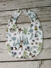 Load image into Gallery viewer, A  bib with a Bambi woodland animal print, featuring deer, rabbits, squirrels, and butterflies among trees and flowers on a white background, laid flat on a rustic wooden surface.
