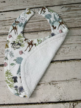 Load image into Gallery viewer, A baby bib with a woodland animal print on the front and a soft white terry cloth backing, displayed on a rustic white-washed wooden surface.
