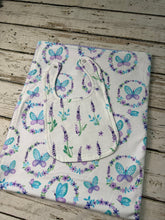 Load image into Gallery viewer, A folded white baby blanket or swaddle with a printed pattern of purple and blue butterflies, lavender sprigs, and floral wreaths, laid flat on a rustic whitewashed wooden surface.
