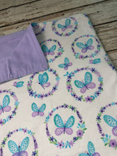 Load image into Gallery viewer, A folded fabric item, likely a blanket or throw, featuring a cream background with a repeating pattern of purple and teal butterflies inside floral wreaths. The reverse side is shown folded over in light purple/lavender, placed on a rustic wooden surface.
