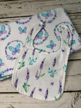 Load image into Gallery viewer, A white fabric baby blanket and bib with a purple, blue, and green floral and butterfly print, displayed on a rustic whitewashed wooden surface.
