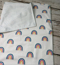 Load image into Gallery viewer, A folded baby blanket, featuring a repeating rainbow pattern in blue, pink, yellow, and orange, with a second folded section showing a tiny blue polka dot pattern on a white background, laid flat on a rustic gray wooden plank surface.

